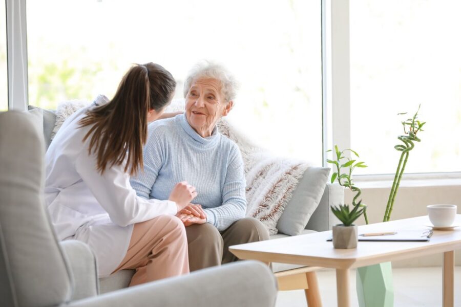 Image alt text Home health clinician holding hands with an older woman on her living room sofa, illustrating home-based care under CMS-0057-F
