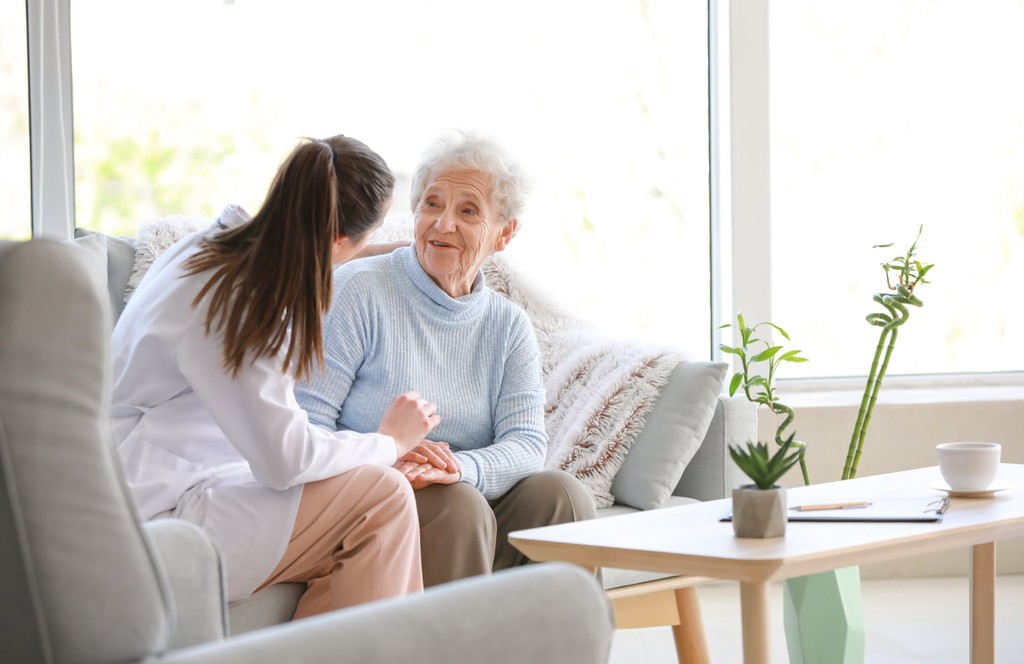 Image alt text Home health clinician holding hands with an older woman on her living room sofa, illustrating home-based care under CMS-0057-F